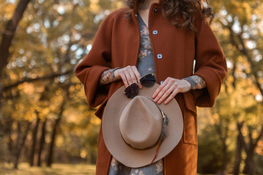 Stylish woman walking in a park wearing a warm chocolate brown coat, holding a hat and sunglasses, fall fashion trend