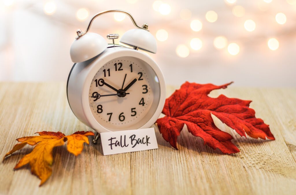 Vintage white alarm clock beside autumn leaves on a wooden table, symbolizing daylight saving time ending and clocks falling back in November.