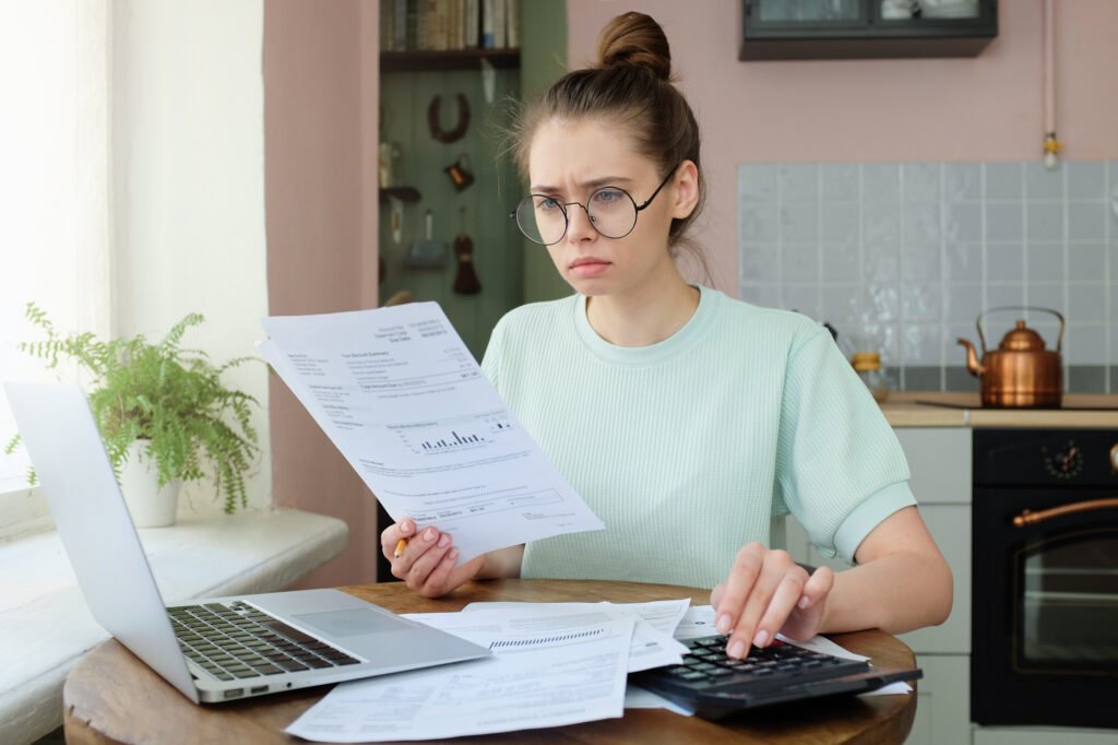 Stressed woman reviewing bills, a calculator, and a laptop at her kitchen table while recalculating her student loan budget